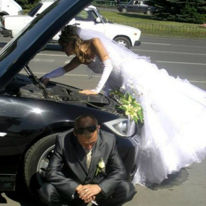 Photo: New bride in her wedding dress repairing her automobile while husband sits on the curb.
